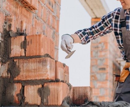 Muncitor aplicând mortar între cărămizi pe un șantier de construcții, unde prezența unui diriginte de santier asigură calitatea execuției