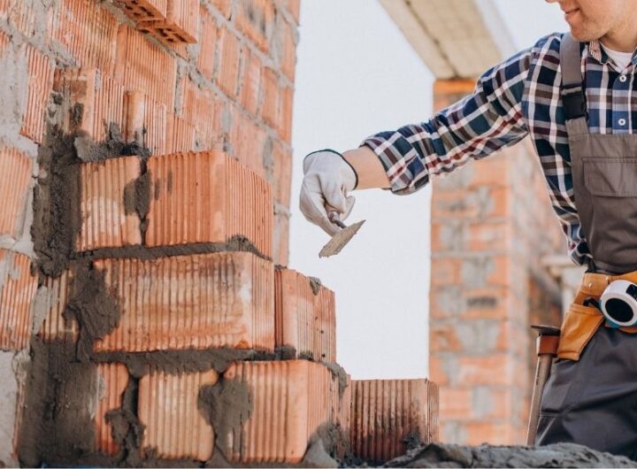 Muncitor aplicând mortar între cărămizi pe un șantier de construcții, unde prezența unui diriginte de santier asigură calitatea execuției